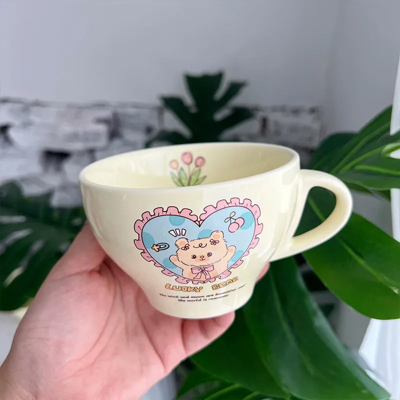 A hand holds a cream-colored ceramic cup featuring a cute cartoon bear character inside a pink heart frame, with green leaves and a blurred indoor background.