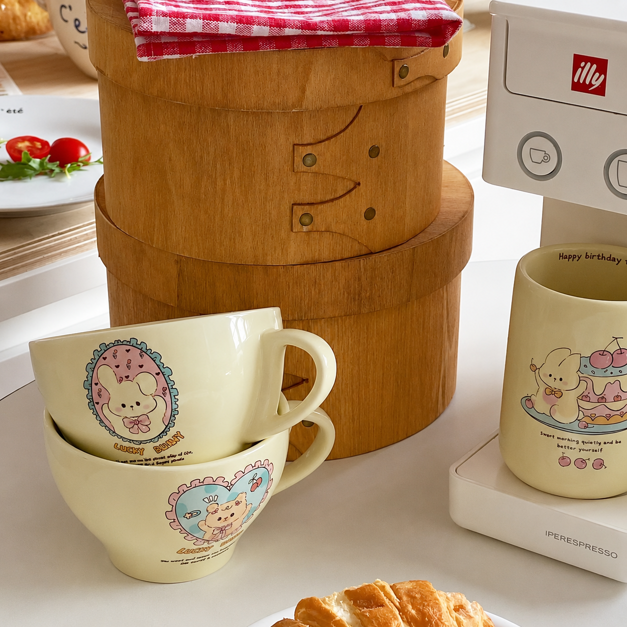 A styled tabletop scene showing a set of cream-colored ceramic mugs and cups with pastel cartoon bunny illustrations, alongside a wooden steamer basket and an Illy espresso machine. The mugs feature pink, blue, and purple bunny-themed designs with small text.