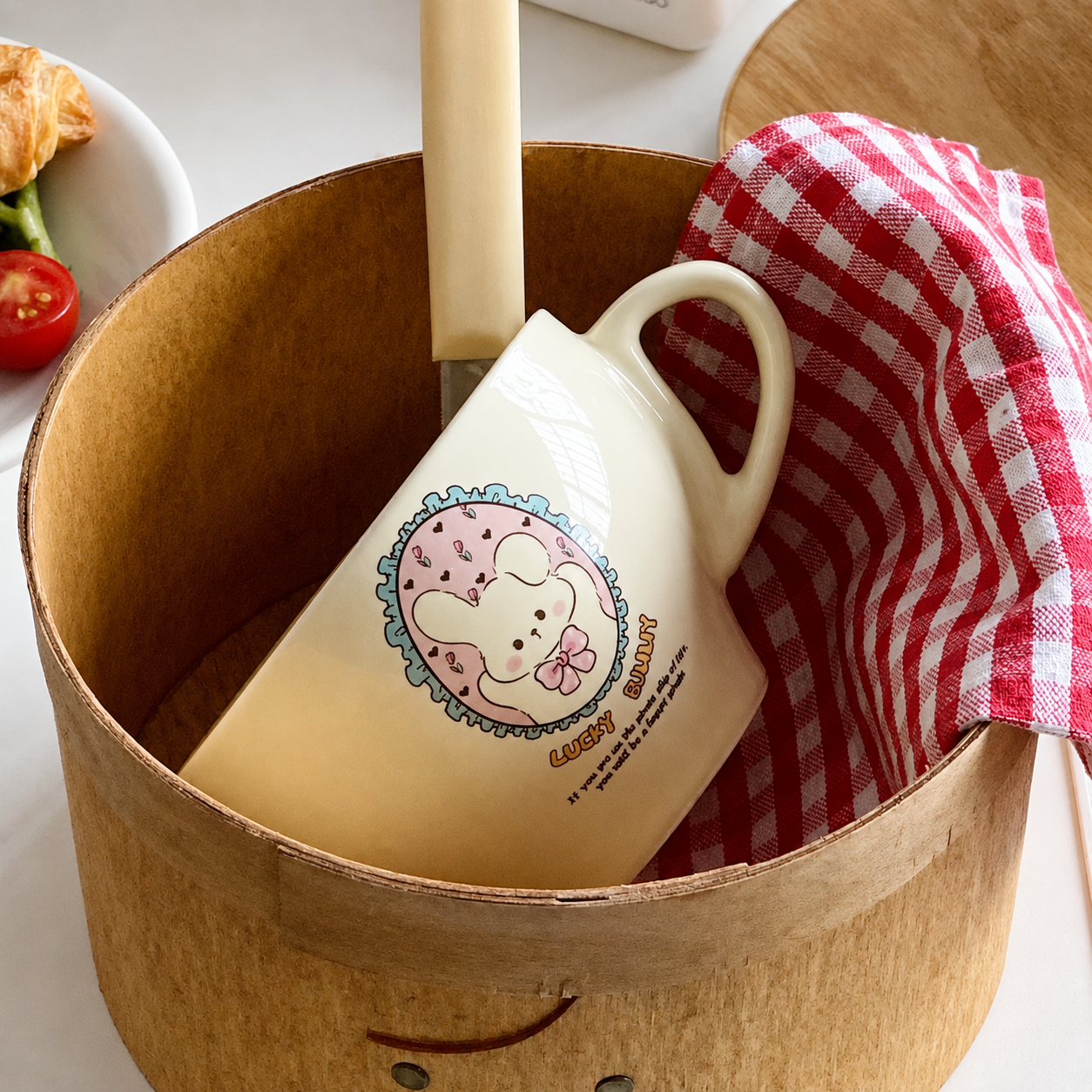 A cream-colored ceramic mug with a pink cartoon bunny illustration and the text 'Cute bunny' inside a round wooden container, alongside a red-and-white checkered cloth and a wooden spoon.