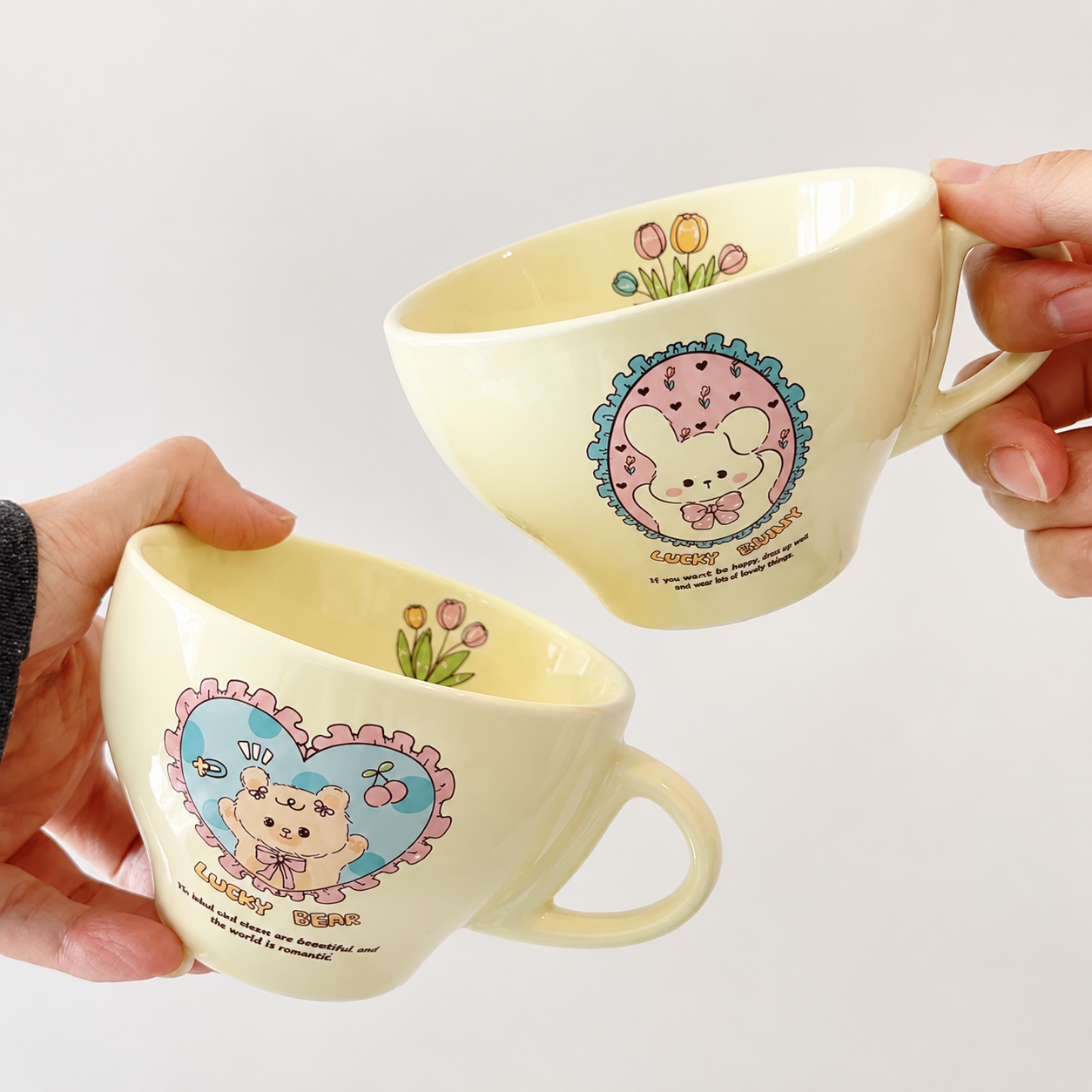 Two cream-colored ceramic mugs with cute cartoon animal illustrations, held by hands against a plain white background.