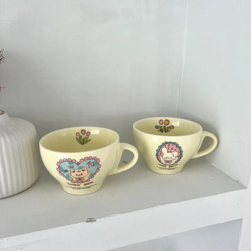 Two pale yellow ceramic cups with cute cartoon animal illustrations, displayed on a white shelf next to a white ribbed vase.