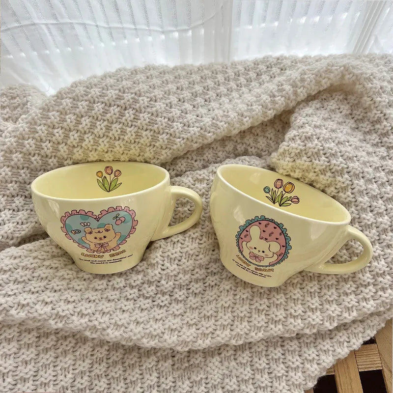 Two pale yellow ceramic cups with cute cartoon bear illustrations and floral designs, placed on a beige knitted blanket in front of a white curtain.