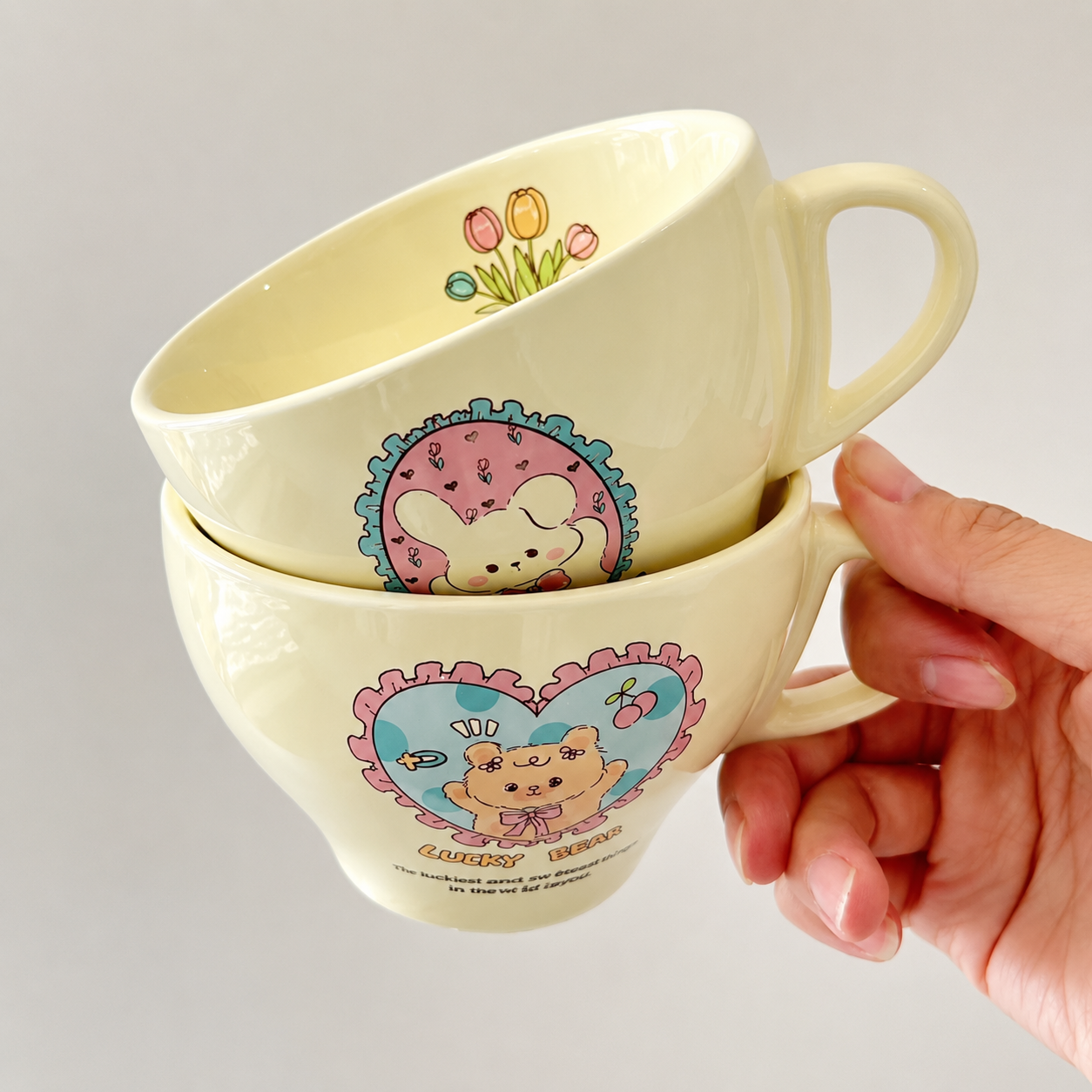 Three stacked cream-colored ceramic mugs with colorful cartoon bear illustrations and floral designs, held by a hand against a plain light gray background.