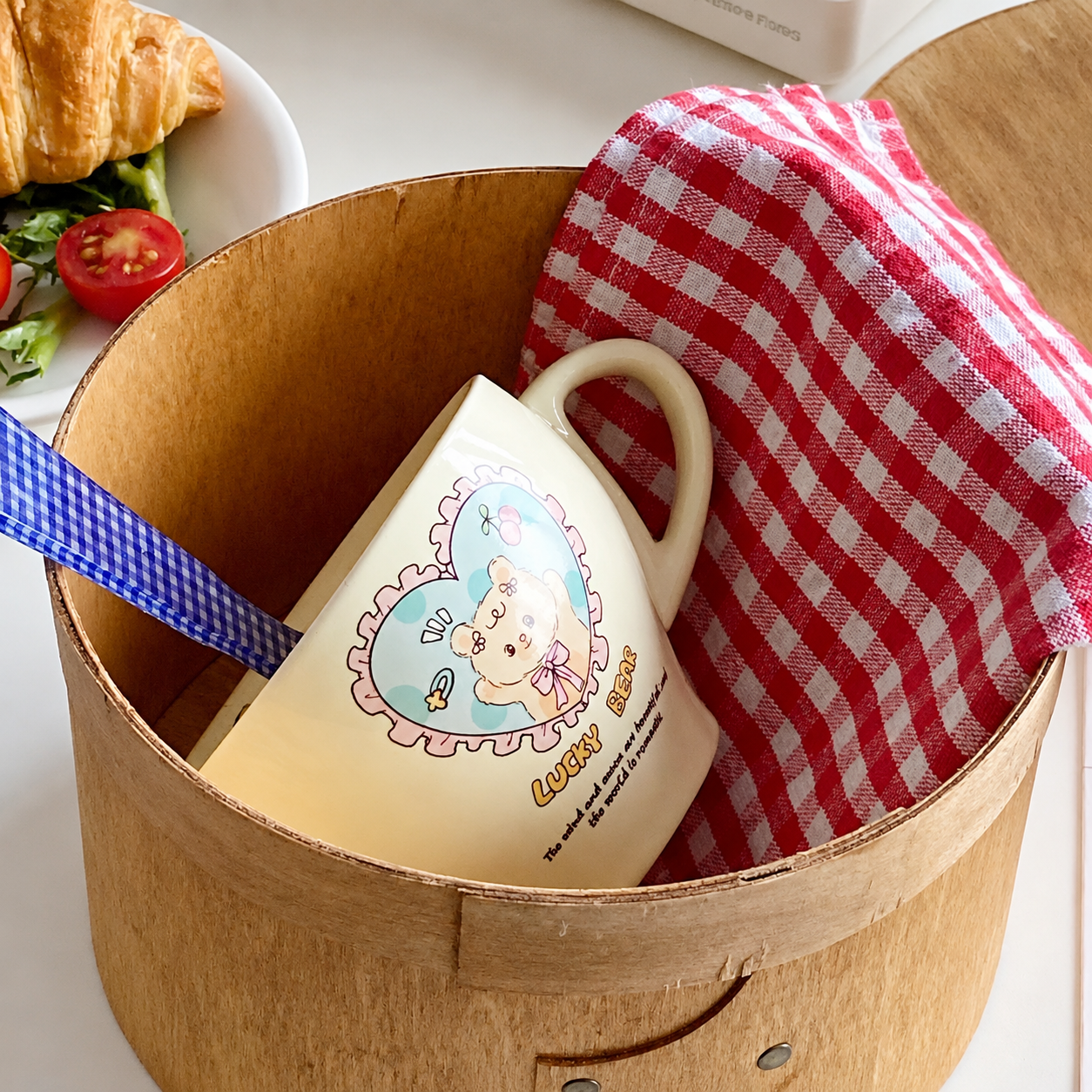 A cream-colored ceramic mug with a cartoon bunny and the text 'LUCKY BUNNY' inside a round wooden box, alongside a red-and-white checkered cloth and a blue gingham strap.