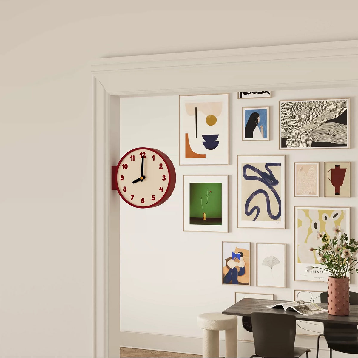 A styled interior scene showing a white wall with a gallery wall of framed abstract art prints, a round red clock, and part of a dining table with chairs and a vase of flowers.