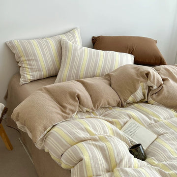 A cozy bed styled with a beige and white striped bedding set, including two striped pillows, a matching duvet cover, and solid beige pillows, set against a plain white wall.