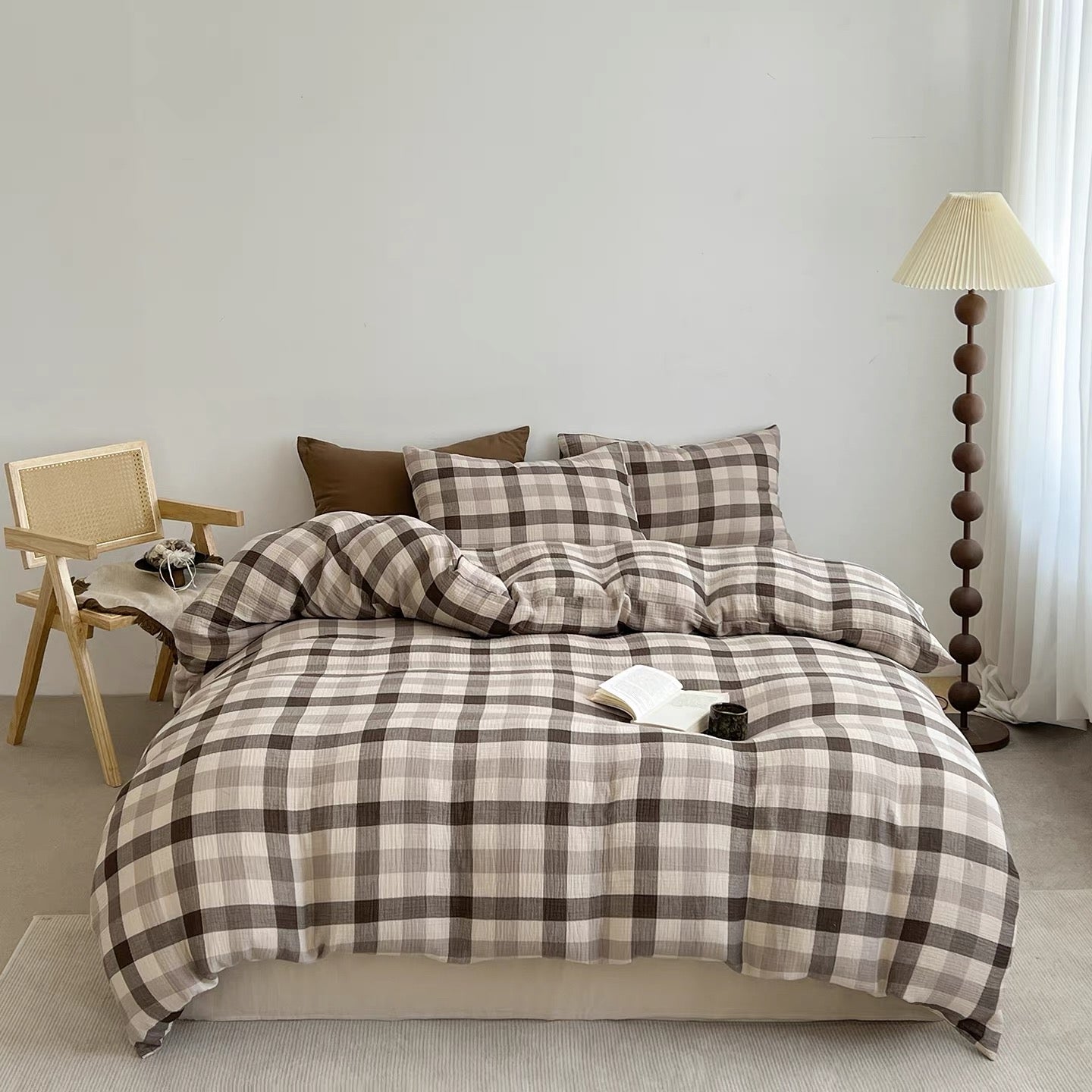 A bed dressed in a beige and brown plaid bedding set, styled in a minimalist bedroom with a wooden chair, floor lamp, and white curtain.