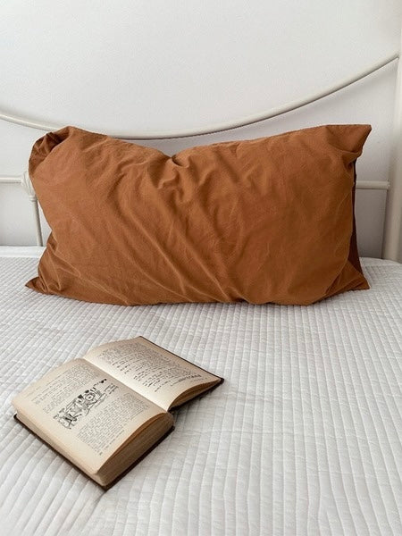 A caramel brown pillowcase rests upright on white bedding with an open book in front.
