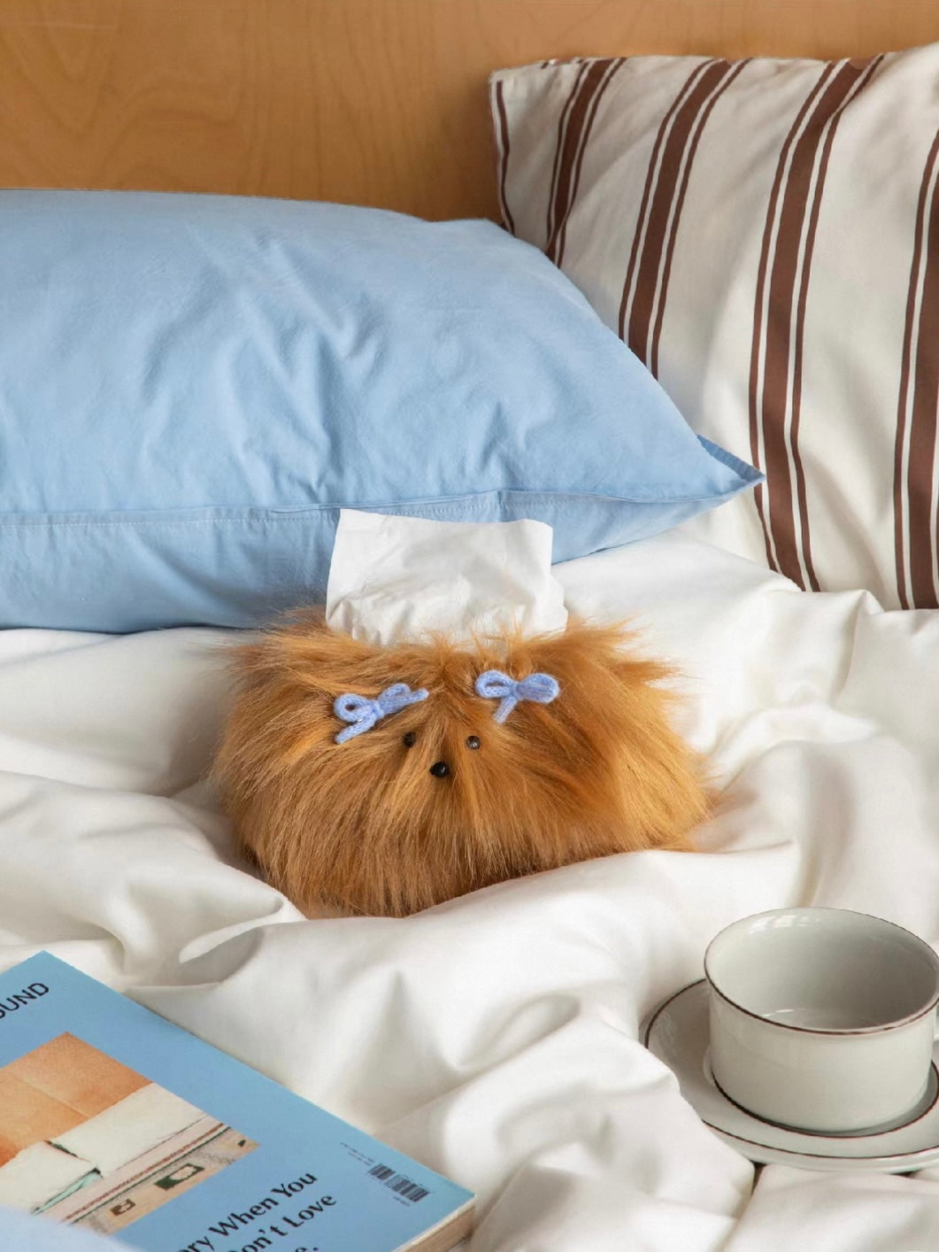 Caramel dog face tissue cover beside a cup and saucer on white bedding, with blue pillows behind it.
