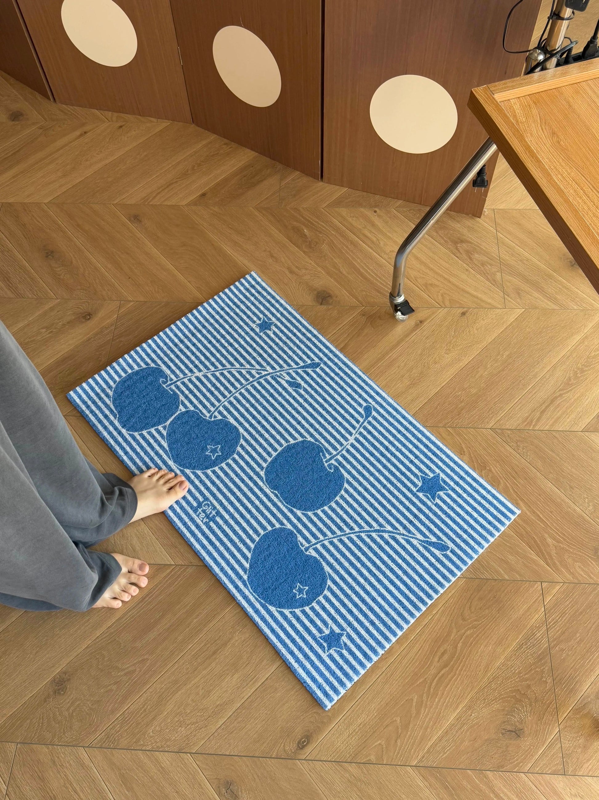 A blue and white striped rectangular mat with a cherry pattern lies on a light wood herringbone floor. A person's bare foot and gray pants are visible at the left edge, and part of a wooden table and cabinet is visible in the room setting.