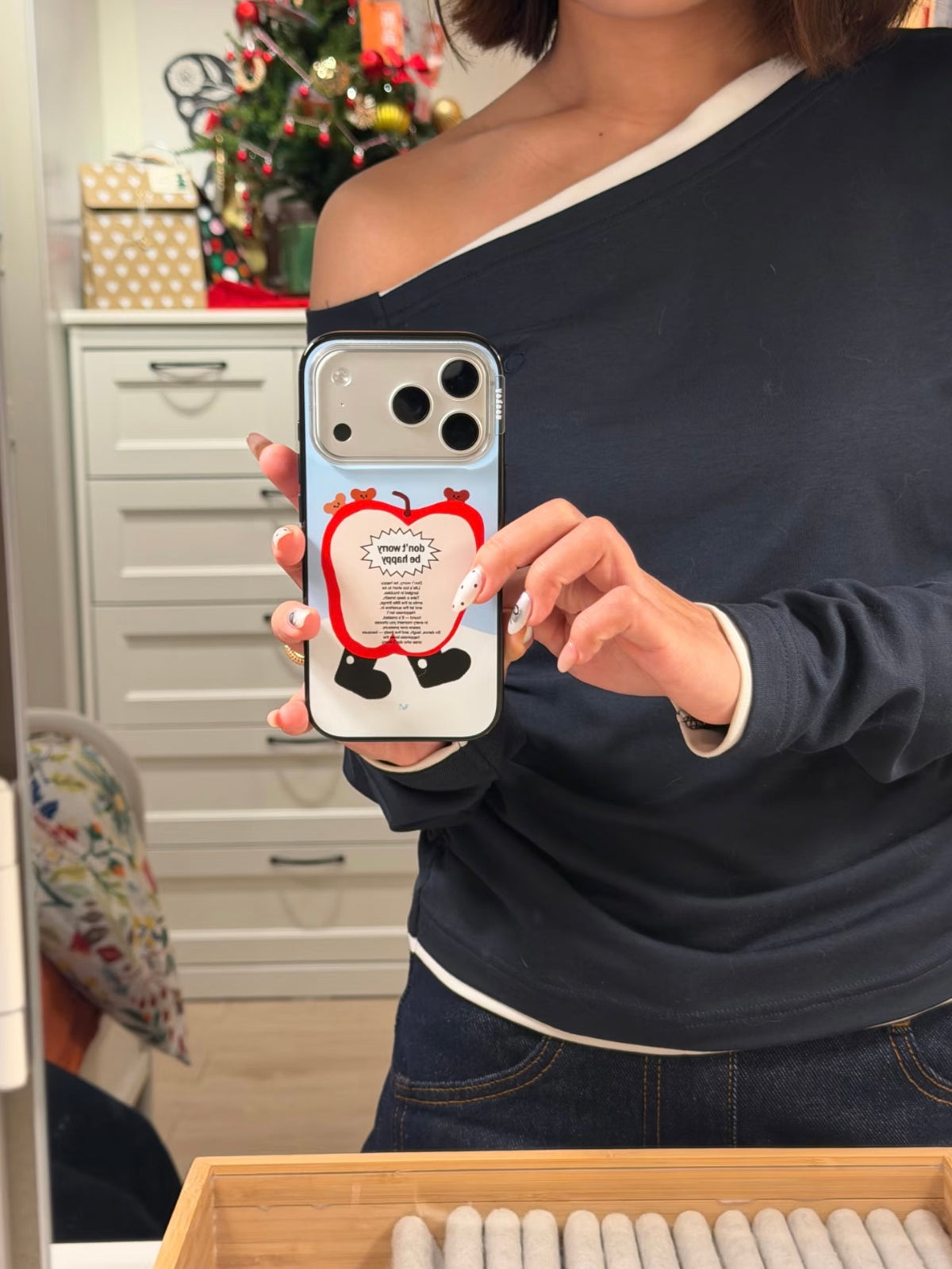 A blue cartoon phone case shows a red apple character and tiny bears as a person takes a mirror selfie.