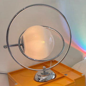 A modern table lamp with a spherical frosted white glass shade and a chrome metal frame, resting on an orange side table against a light wall.