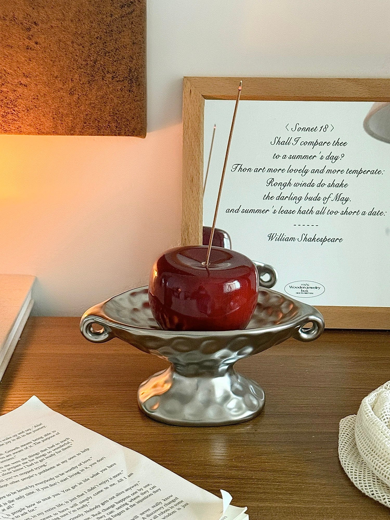 A red apple-shaped candle with a wooden wick sits in a silver pedestal dish on a wooden table, styled with a framed Shakespeare quote print and warm ambient lighting.