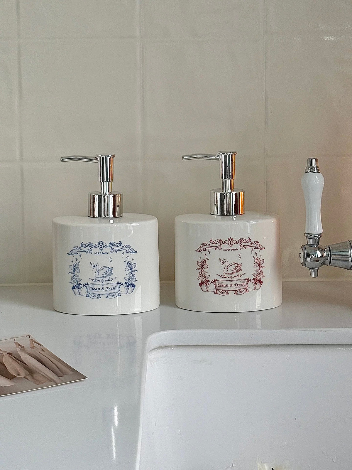 Two ceramic soap dispensers stand beside a faucet, with blue and red swan artwork on white bottles.