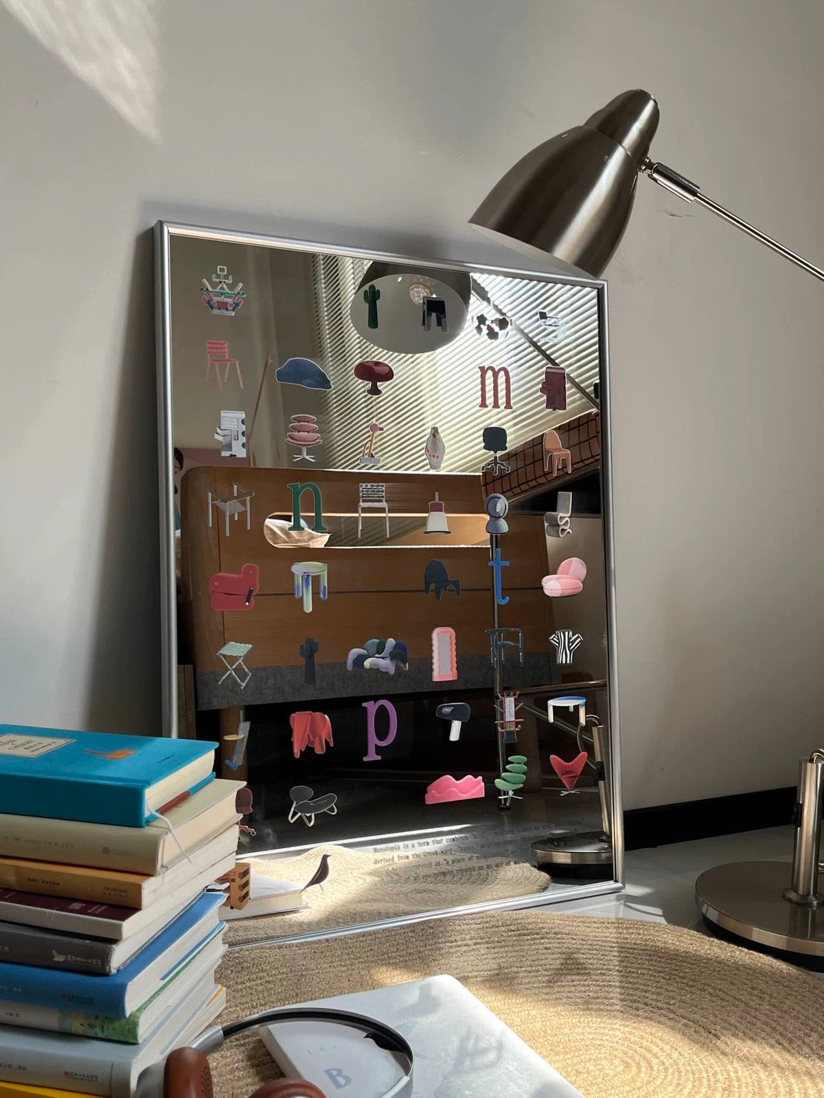 A tall rectangular mirror with a silver frame leans against a wall in a styled room. The mirror surface is covered with colorful, small furniture illustrations and letters.