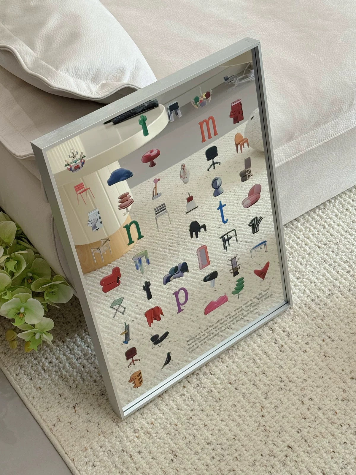 A framed furniture-themed art print leaning against a sofa on a textured cream rug. The print features a silver frame and displays various colorful chairs, tables, and seating items arranged in a grid with letters.