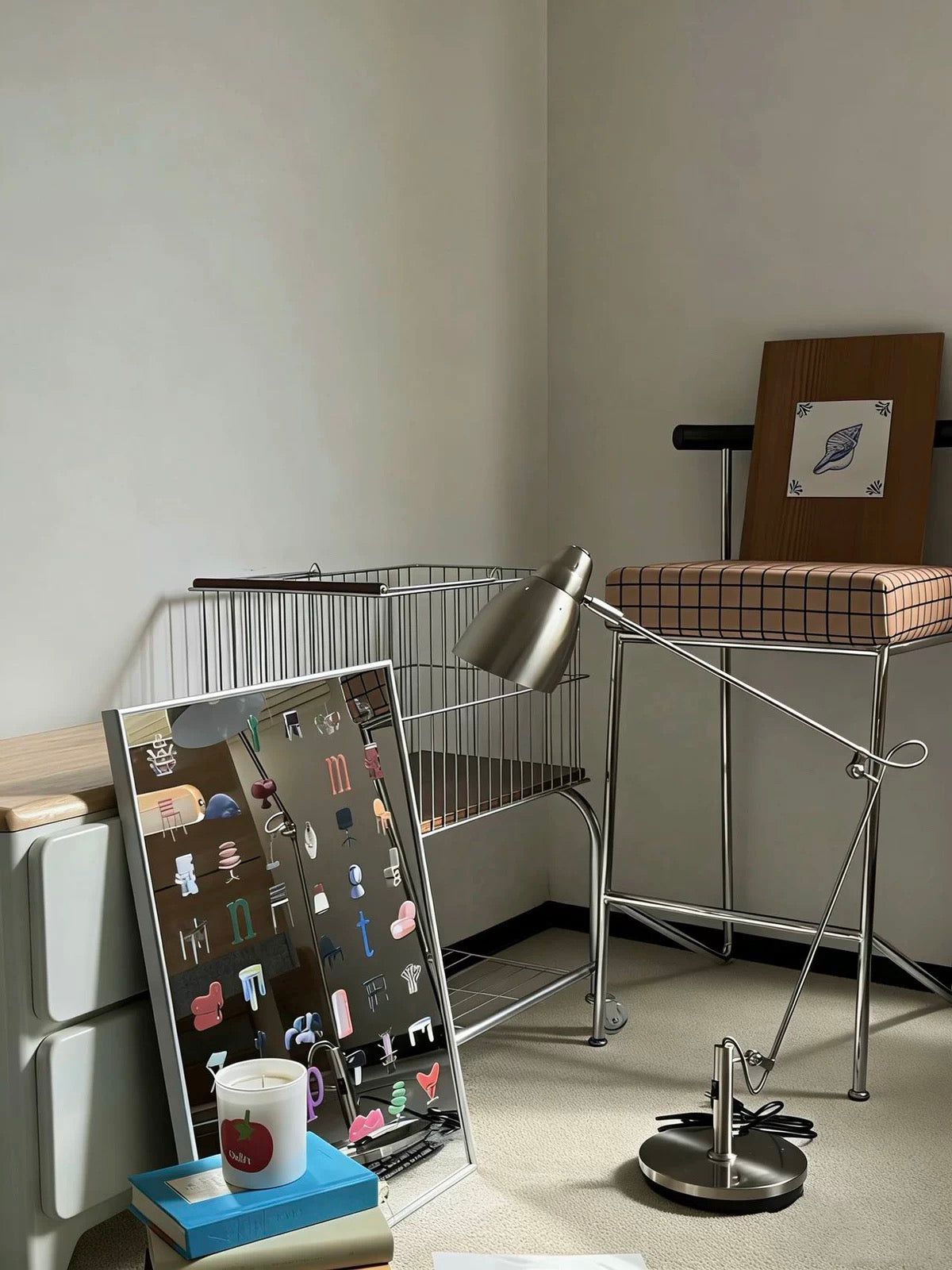 A styled interior corner showing a tall floor lamp with a silver metal arm and round base, next to a metal chair with a plaid cushion, and a leaning mirror on books in a minimalist room.