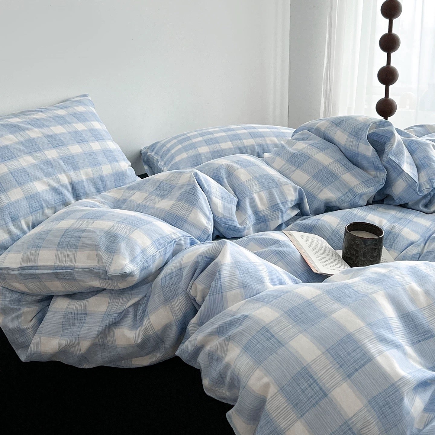 Light blue and white plaid bedding set with matching pillowcases and duvet, styled on a bed in a bright bedroom with a book and cup on top.