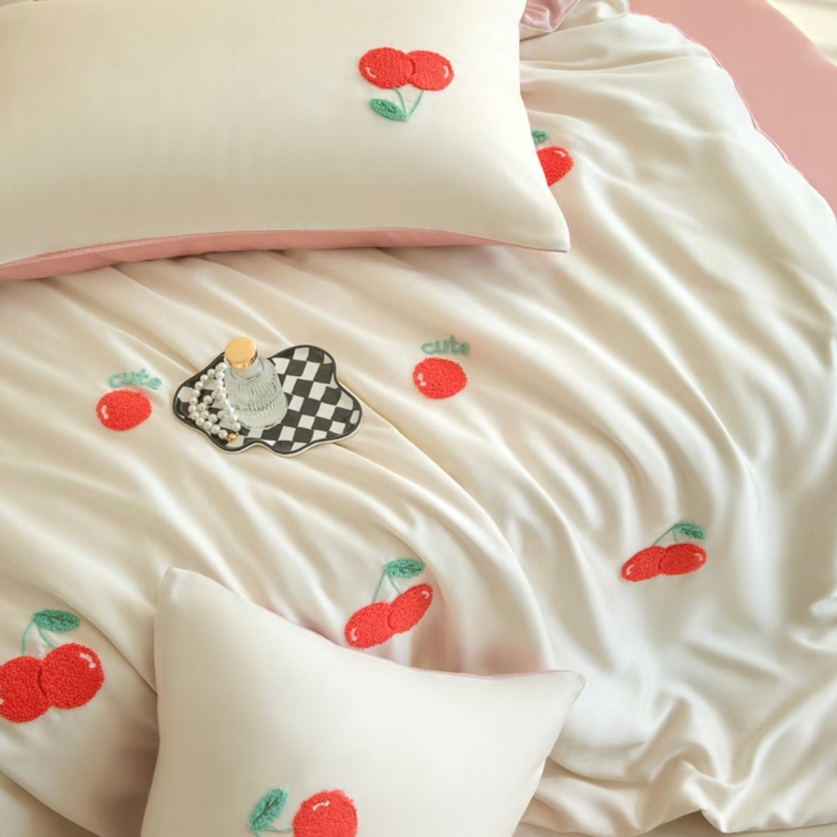 A close-up of a satin bedspread and matching pillowcases in a warm cream color, decorated with embroidered red cherries and green leaves. A decorative tray with pearls and a small bottle is placed on the bedspread.