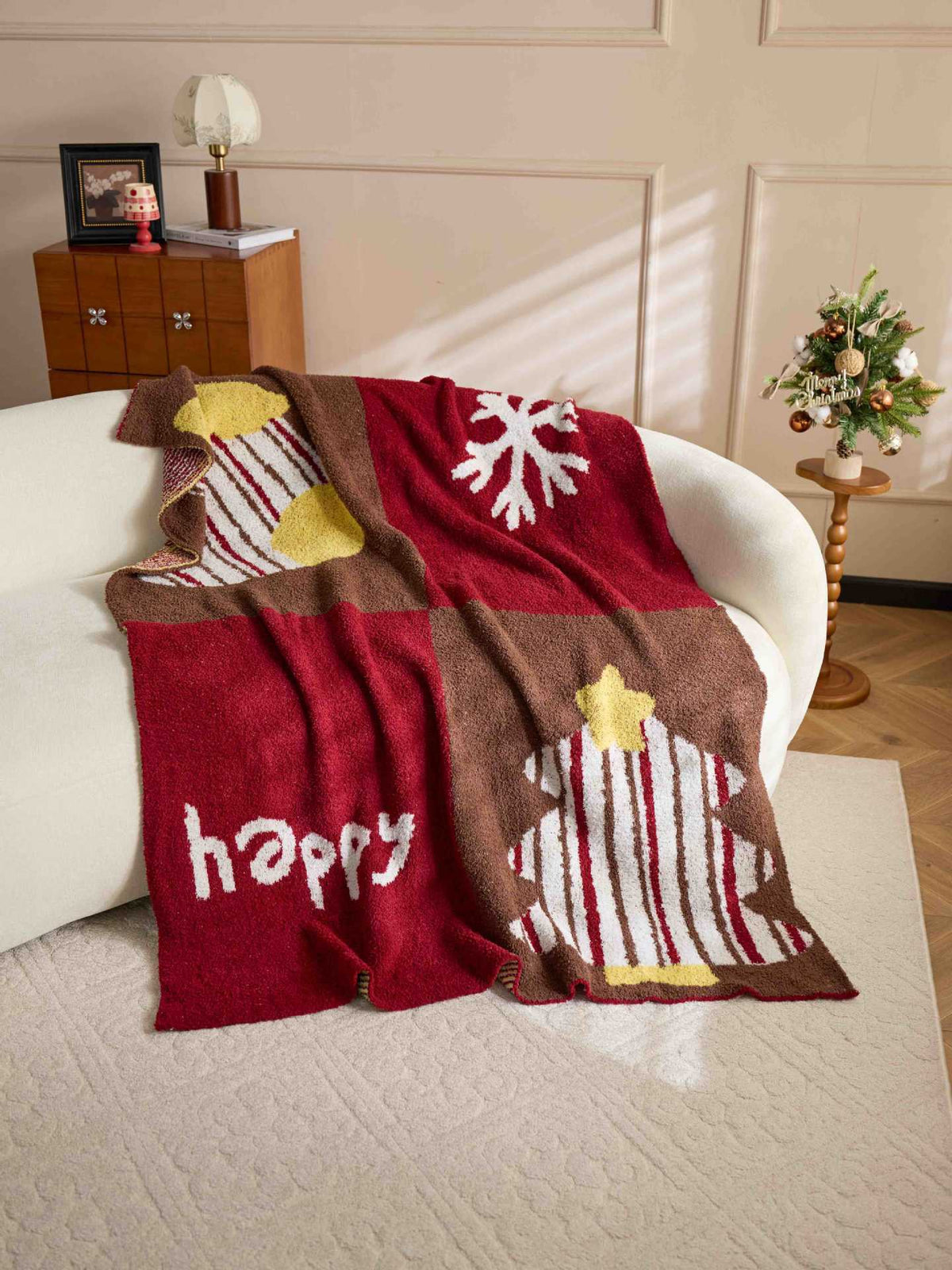 A cozy Christmas-themed knit throw blanket draped over a white armchair in a warmly lit living room. The blanket features a red and brown patchwork design with white snowflakes, striped stockings, a Christmas tree, and the word 'happy' in white.