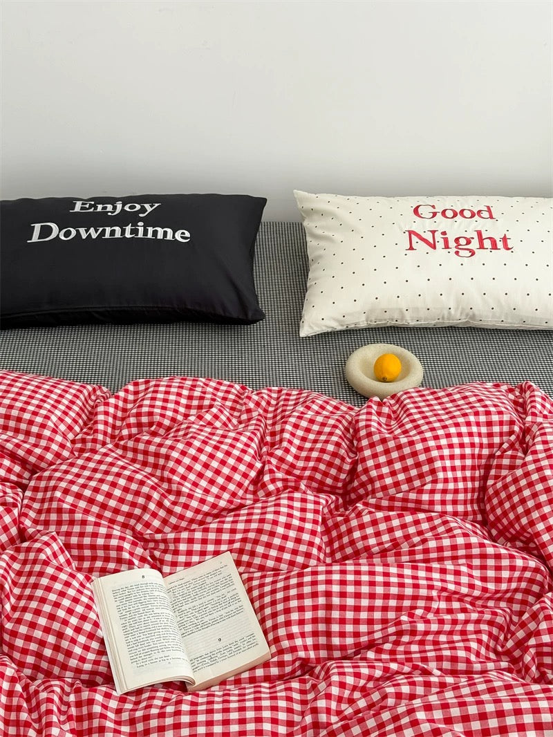 Bed with red checkered duvet, black pillow with 'Enjoy Downtime', white pillow with 'Good Night', and an open book.