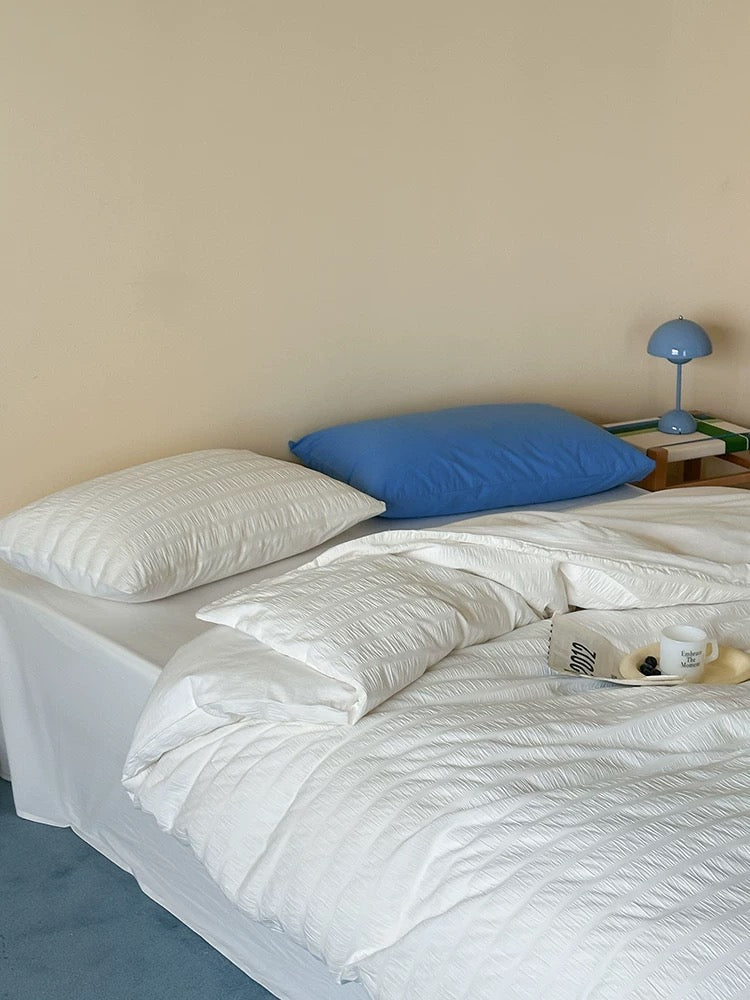 A neatly made bed with white textured bedding and two pillows, one white and one blue, in a minimalist bedroom setting with a beige wall and a small wooden nightstand.