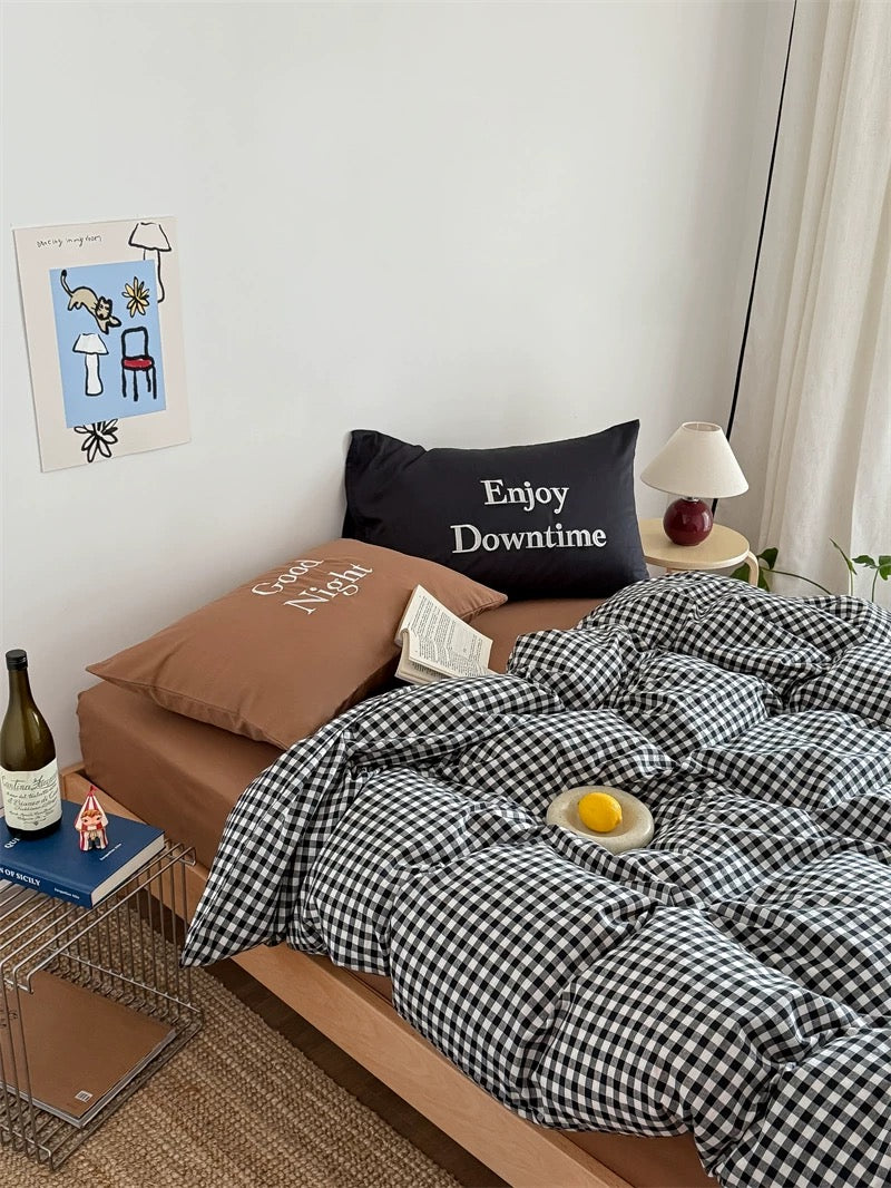 Bedroom with checkered duvet, pillows, and a small table with a bottle and book.