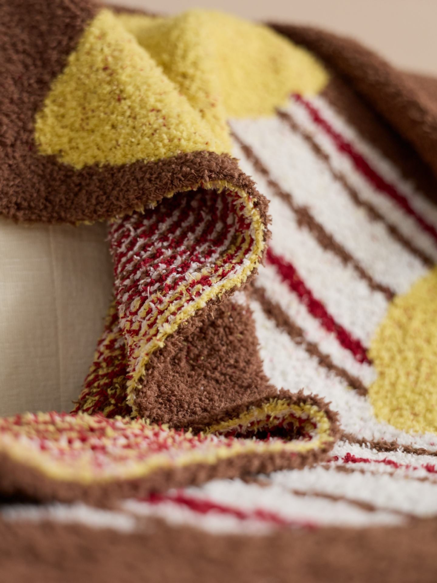 A close-up of a plush, textured throw blanket with a geometric pattern in brown, yellow, white, and red, draped over a light-colored sofa.