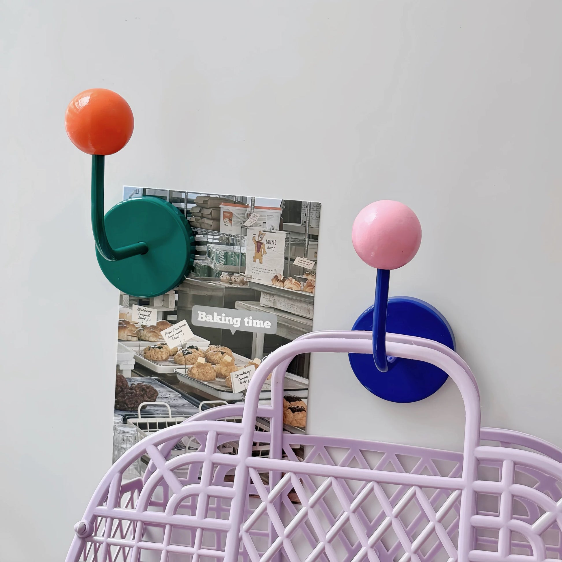 Two wall-mounted hook holders with round bases and spherical tops, shown in teal, orange, pink, and blue. One holder holds a baking shop poster, and the other is attached to a lavender wire basket against a plain light wall.