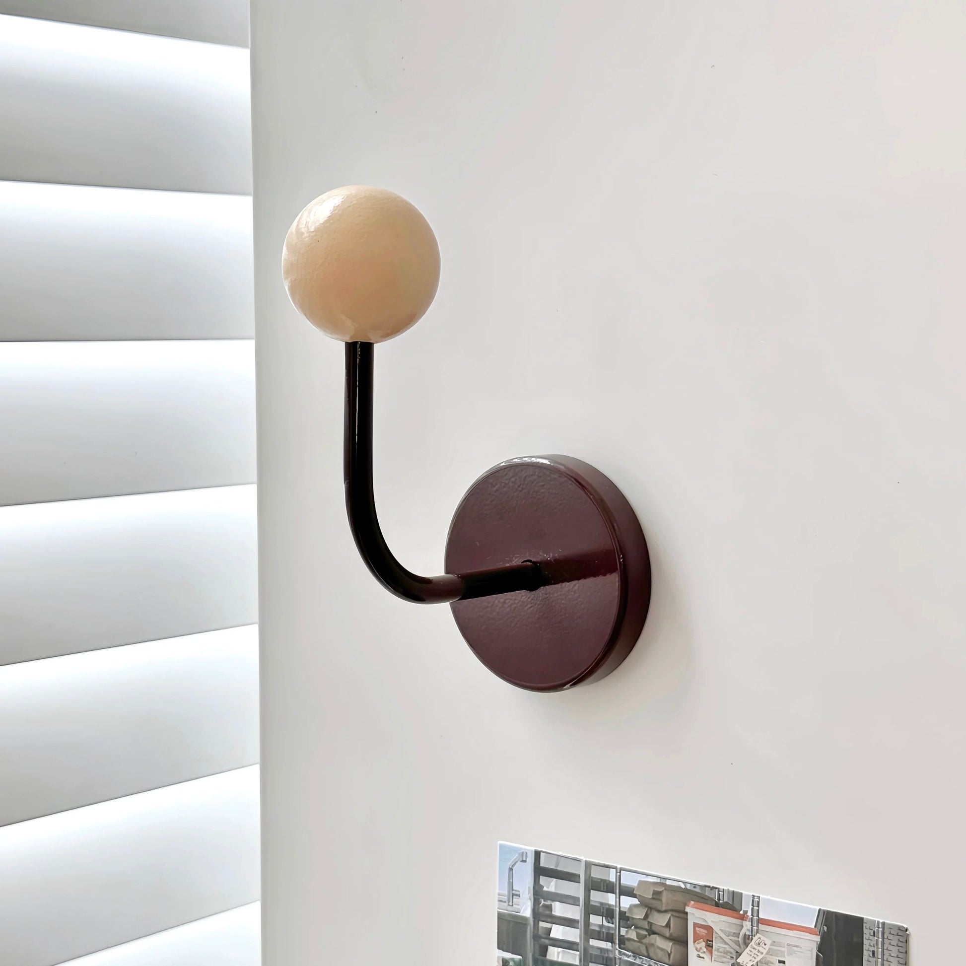 A modern wall-mounted coat hook with a spherical cream-colored top and a curved black stem attached to a round dark reddish-brown wall plate, shown mounted on a white wall next to window blinds.