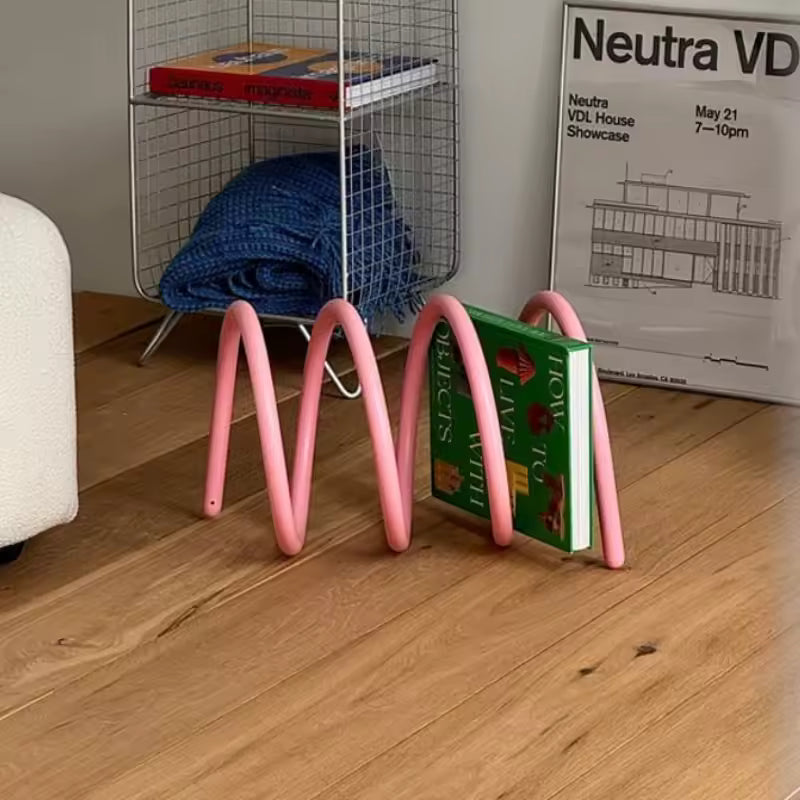 Pink metal bookend holding a green book on a wooden floor with a wire basket in the background.