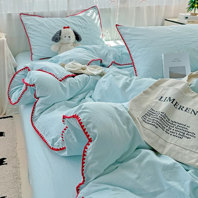 A light blue bedding set with red zigzag trim, styled on a bed with matching pillows, a plush dog toy, a small bow, and a cream tote bag in a bright bedroom setting.