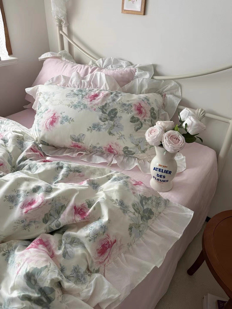 Floral bedding set on a bed with a small vase of flowers and a bottle labeled 'Atelier des' in the foreground.