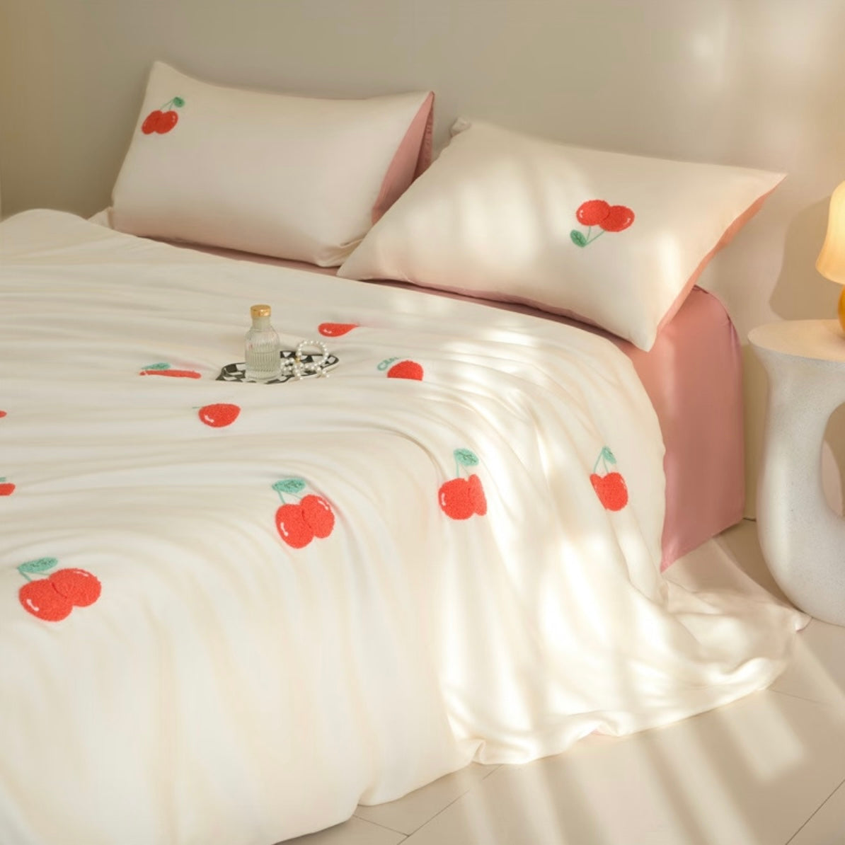 A neatly made bed with white bedding printed with red cherries and green leaves, styled in a warm, softly lit room with pink pillows and a pink bed frame visible.