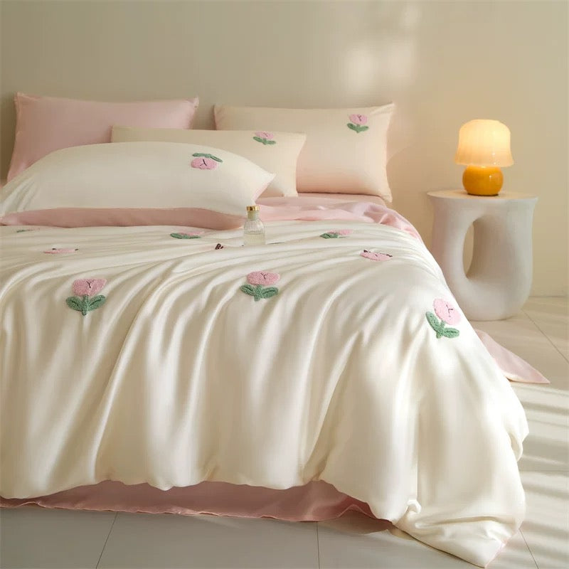 A bed dressed in a soft cream bedding set with pink floral embroidery and a blush pink bed skirt, styled in a warm, softly lit bedroom with matching pillows and a small bedside lamp.