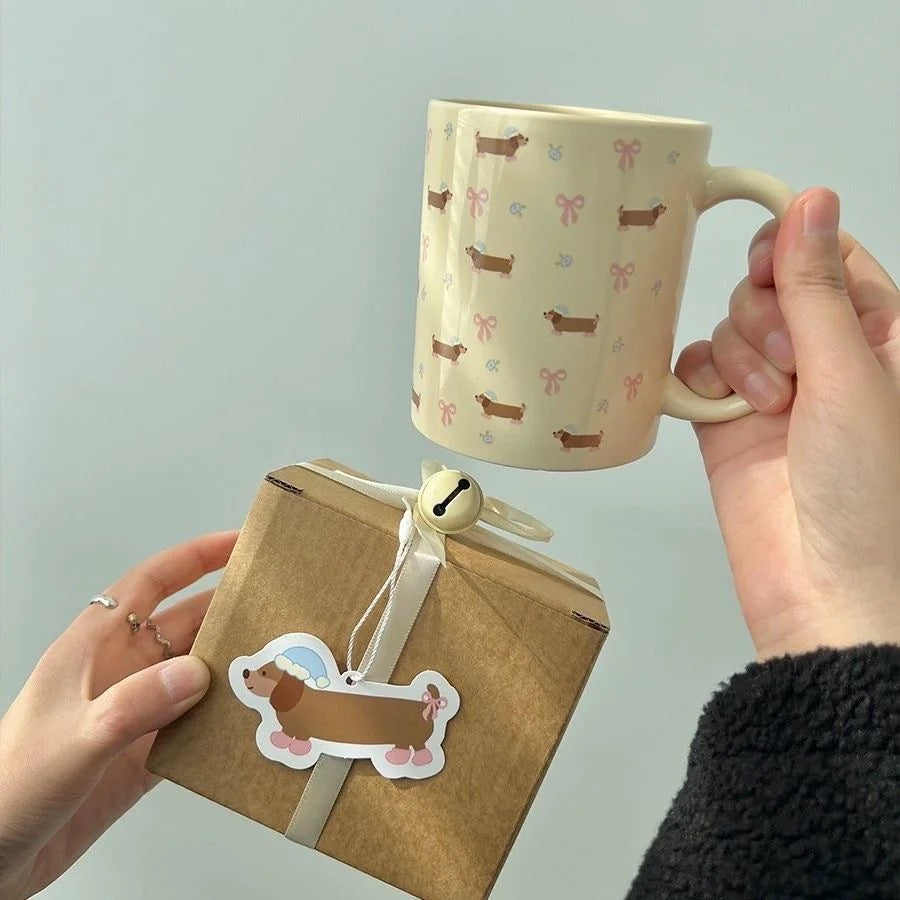 A cream-colored ceramic mug with a dachshund and bow pattern is held by a hand above a small kraft paper gift box tied with a ribbon and a dachshund sticker.