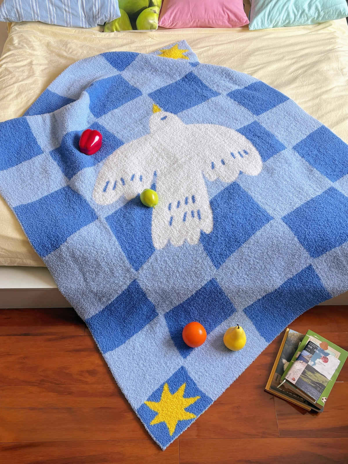 A blue and white checkered plush blanket with a white dove motif and yellow star accents, draped over a bed in a bedroom setting with colorful pillows, wooden floor, and small toys.