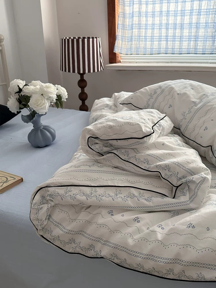 Neatly made bed with floral bedding, lamp, and flowers on a table in a bedroom.