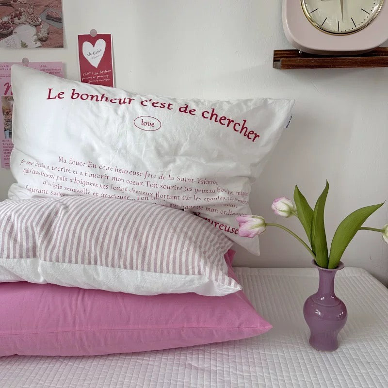 Decorative pillows with French text on a bed with a vase of flowers and clock in the background.