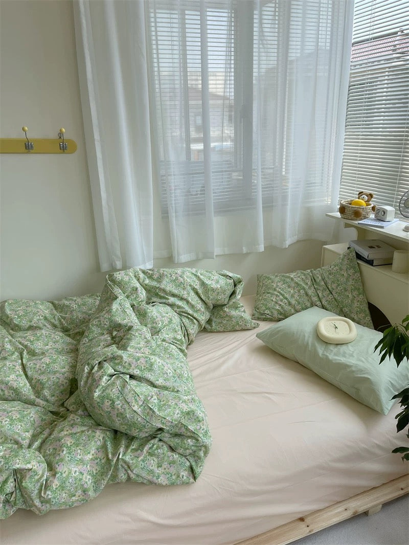 Bed with green floral bedding and pillows in a room with white curtains and a small table.