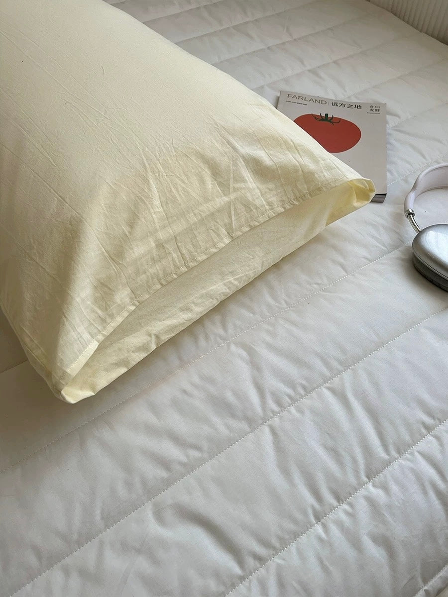 A pale cream pillowcase resting on a white quilted bedspread, with a book and a pair of headphones nearby as styled props.