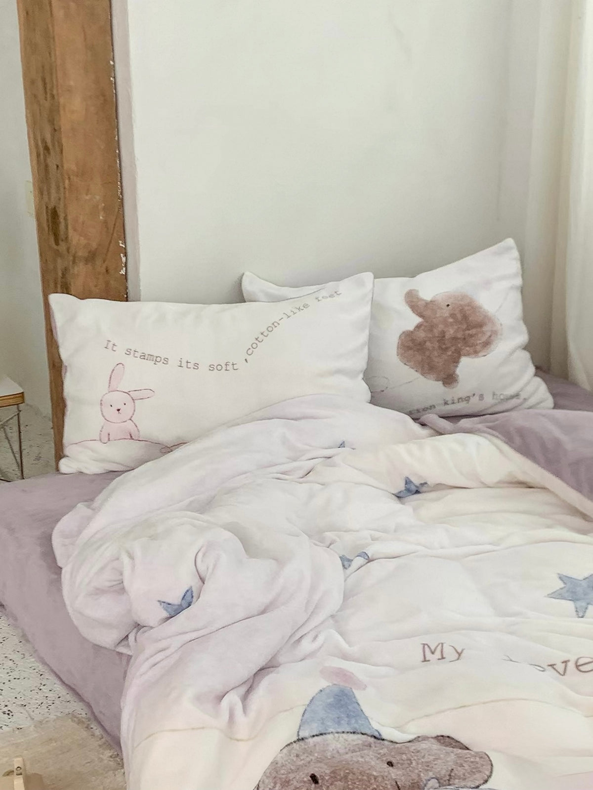 Cream and pink star bedding with elephant and rabbit pillowcases beside a bright curtain.