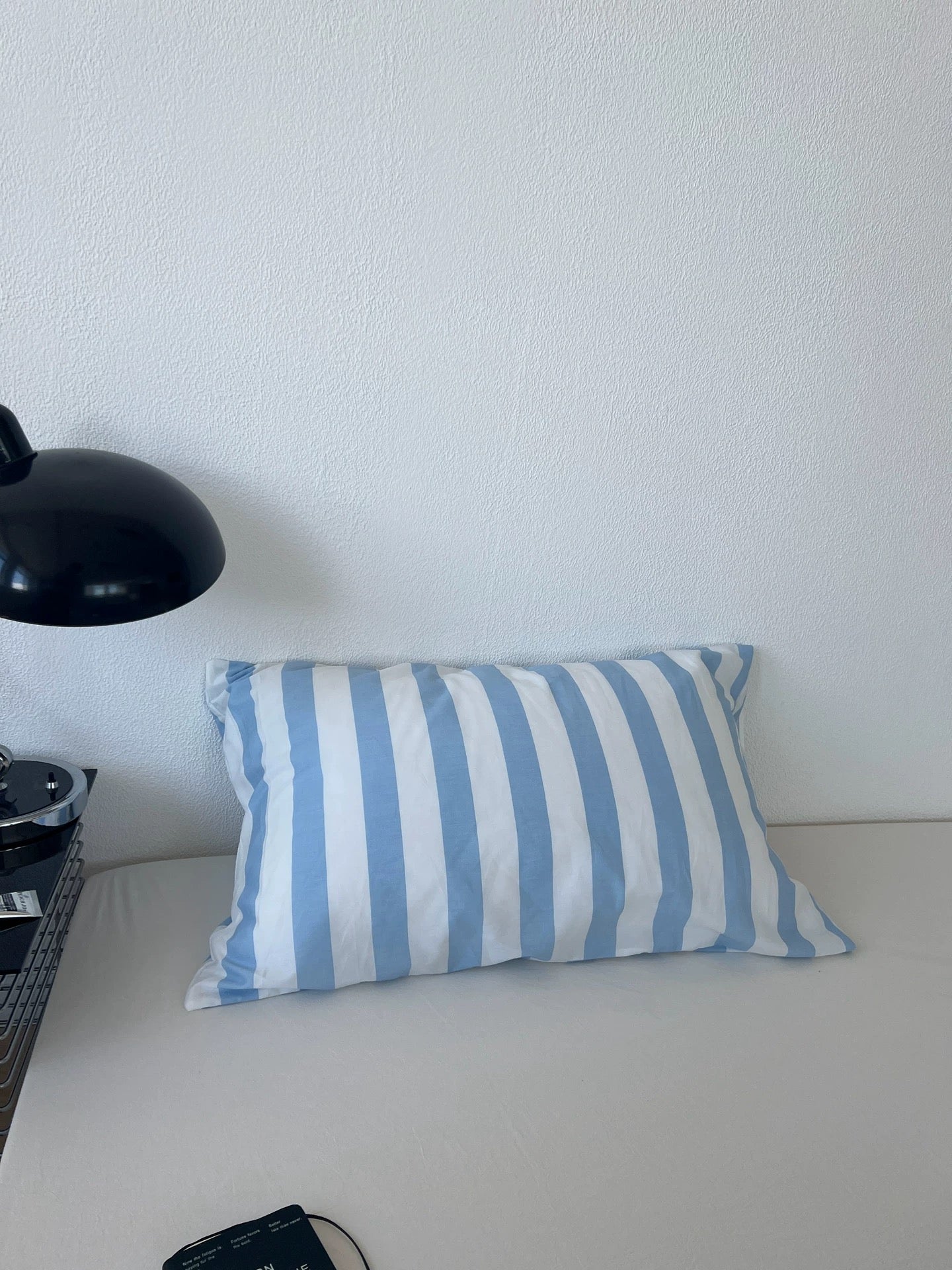 A rectangular throw pillow with vertical light blue and white stripes, resting on a light-colored sofa against a white wall, with a black lamp partially visible on the left.