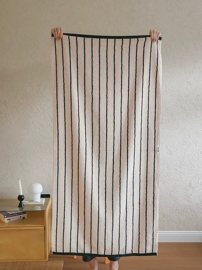 A person holds up a large rectangular textile, likely a towel or blanket, featuring vertical black stripes on a warm cream background. The textile is displayed in a home setting with a textured beige wall, a wooden side table, and a lamp in the background.