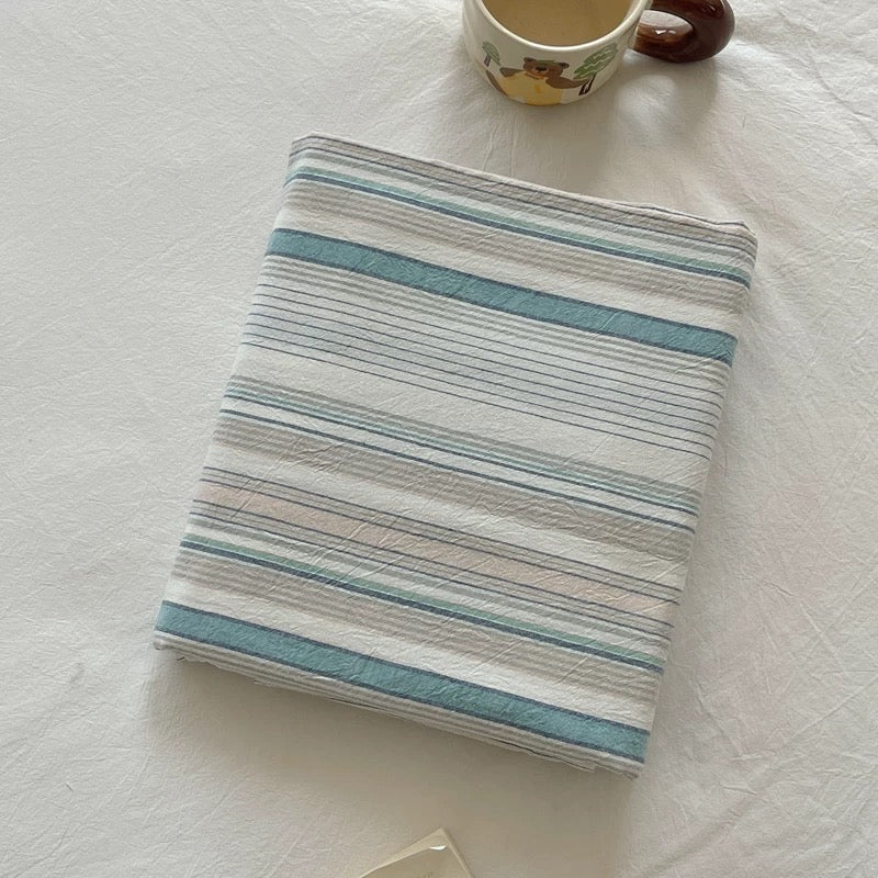 A folded white muslin fabric item, likely a baby blanket or swaddle, featuring horizontal stripes in light blue and thin gray lines, styled on a white textured blanket with a ceramic mug partially visible at the top.
