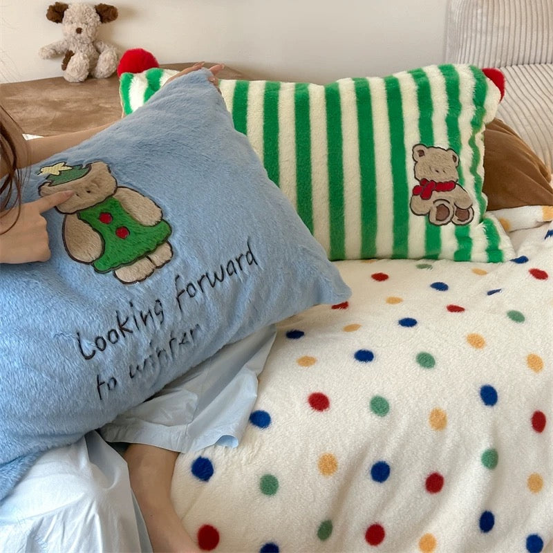 Person holding a blue pillow with a teddy bear design and text, sitting on a bed with colorful polka dot blanket and striped pillow.