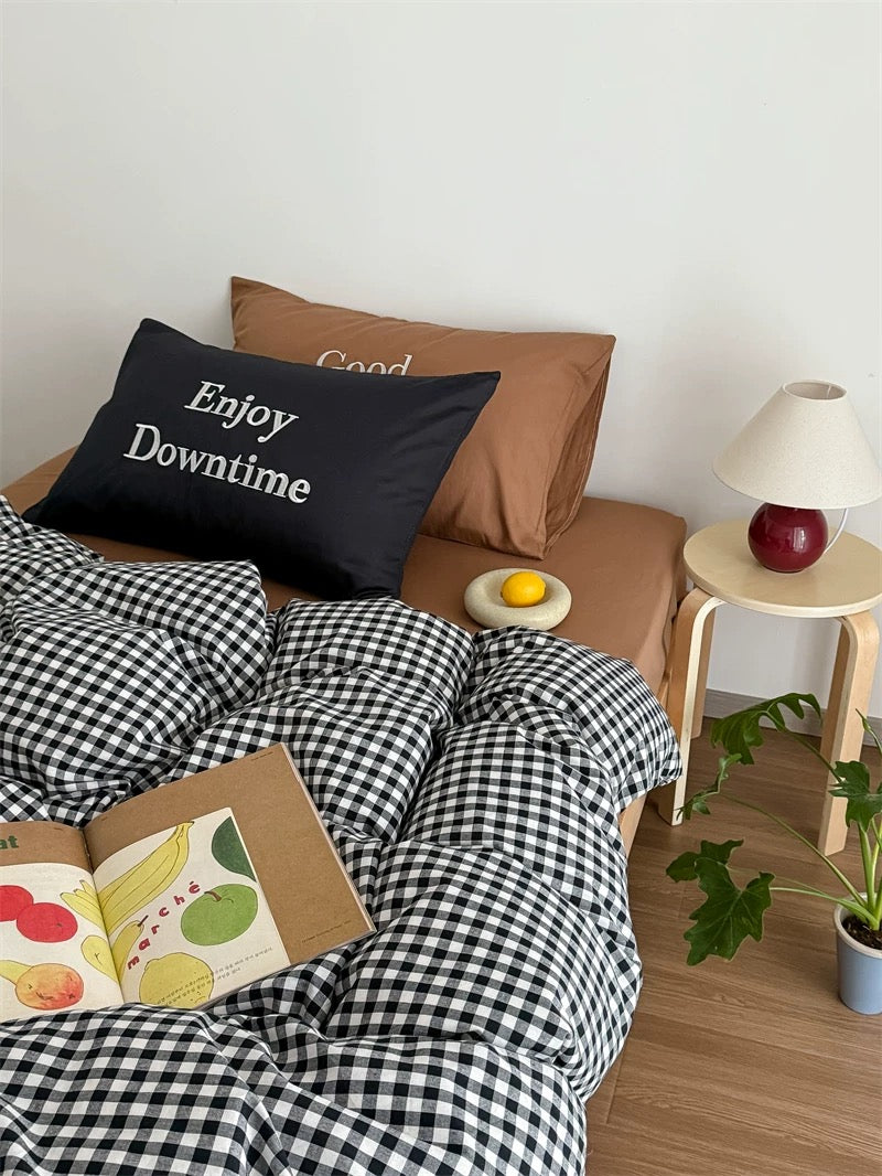 Bed with checkered duvet, pillow with 'Enjoy Downtime' text, and a small table with a lamp and fruit.