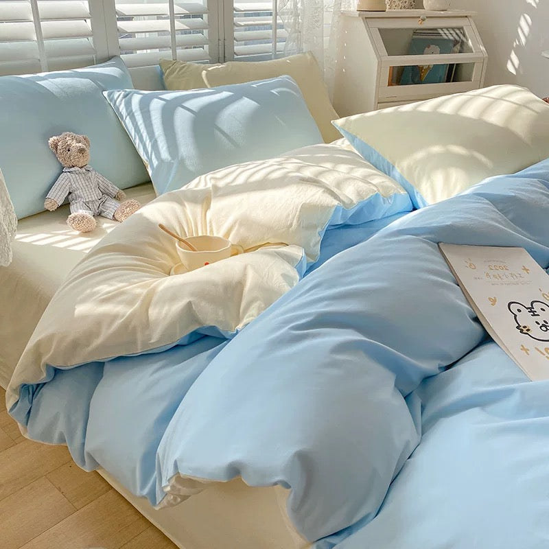 Light blue and beige bedding set on a bed with a teddy bear and book in a bright room.