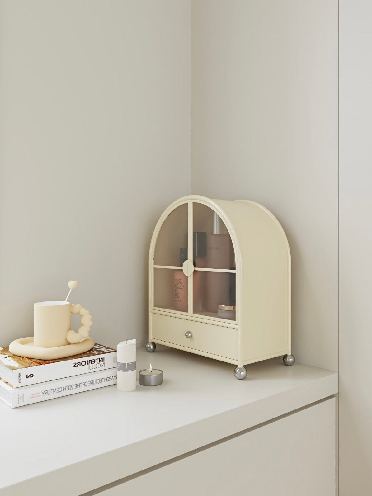 A cream arched cabinet is shown at an angle on a dresser, with silver ball feet and bottles inside.