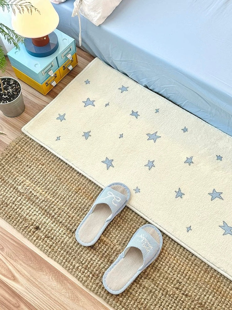 A cream rug with blue stars lies flat beside a bed, with the full near edge and blue slippers visible.